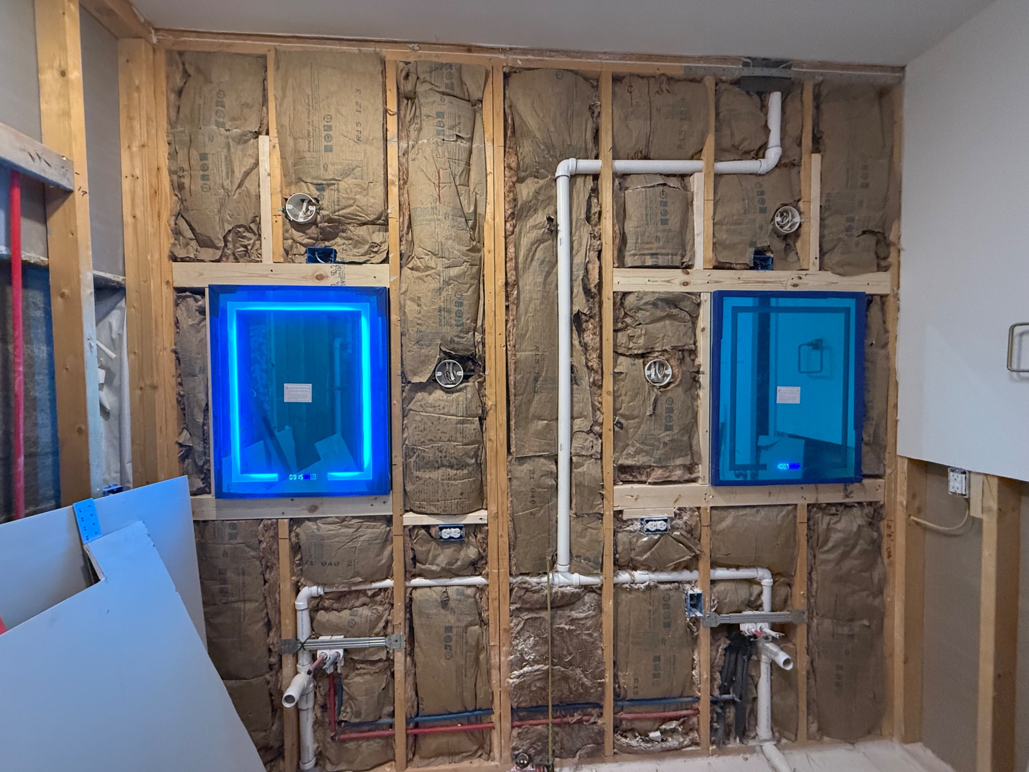 Bathroom wall with plumbing rough-in, insulation, electrical boxes, and illuminated mirrors during construction